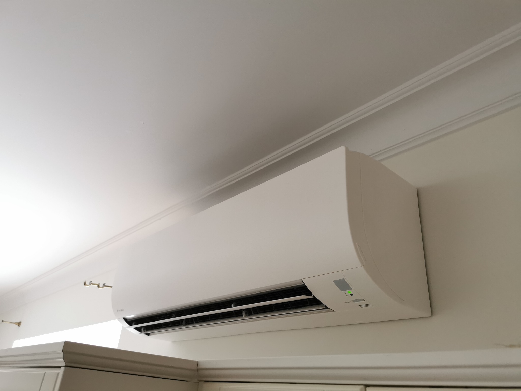 Biała jednostka klimatyzacyjna Daikin zamontowana na ścianie w pomieszczeniu z dekoracyjną listwą przysufitową i widocznymi fragmentami mebli.