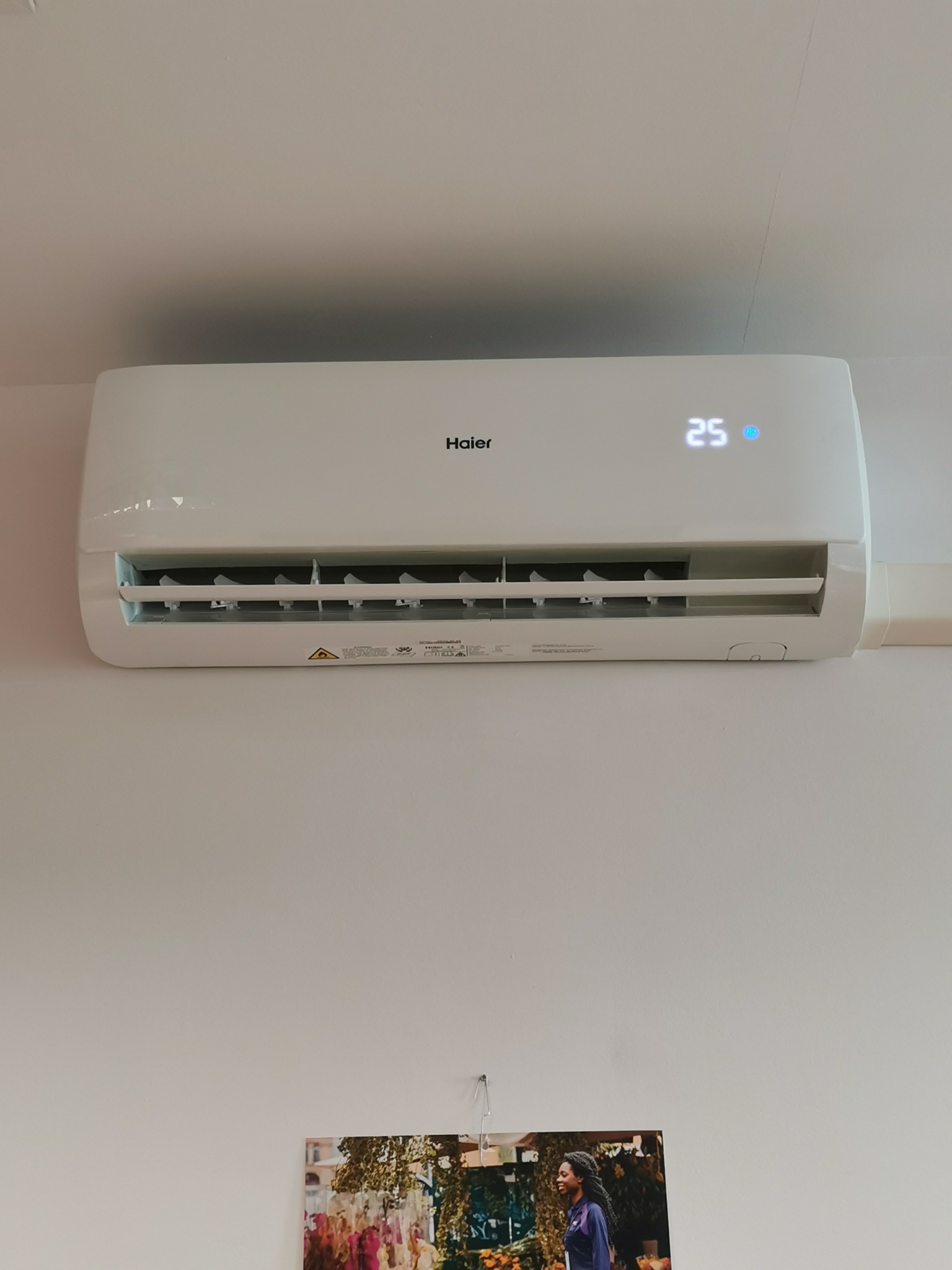 Biała jednostka wewnętrzna klimatyzacji Haier zamontowana na białej ścianie, wyświetlająca temperaturę 25 stopni Celsjusza, z widocznym zdjęciem zawieszonym poniżej.