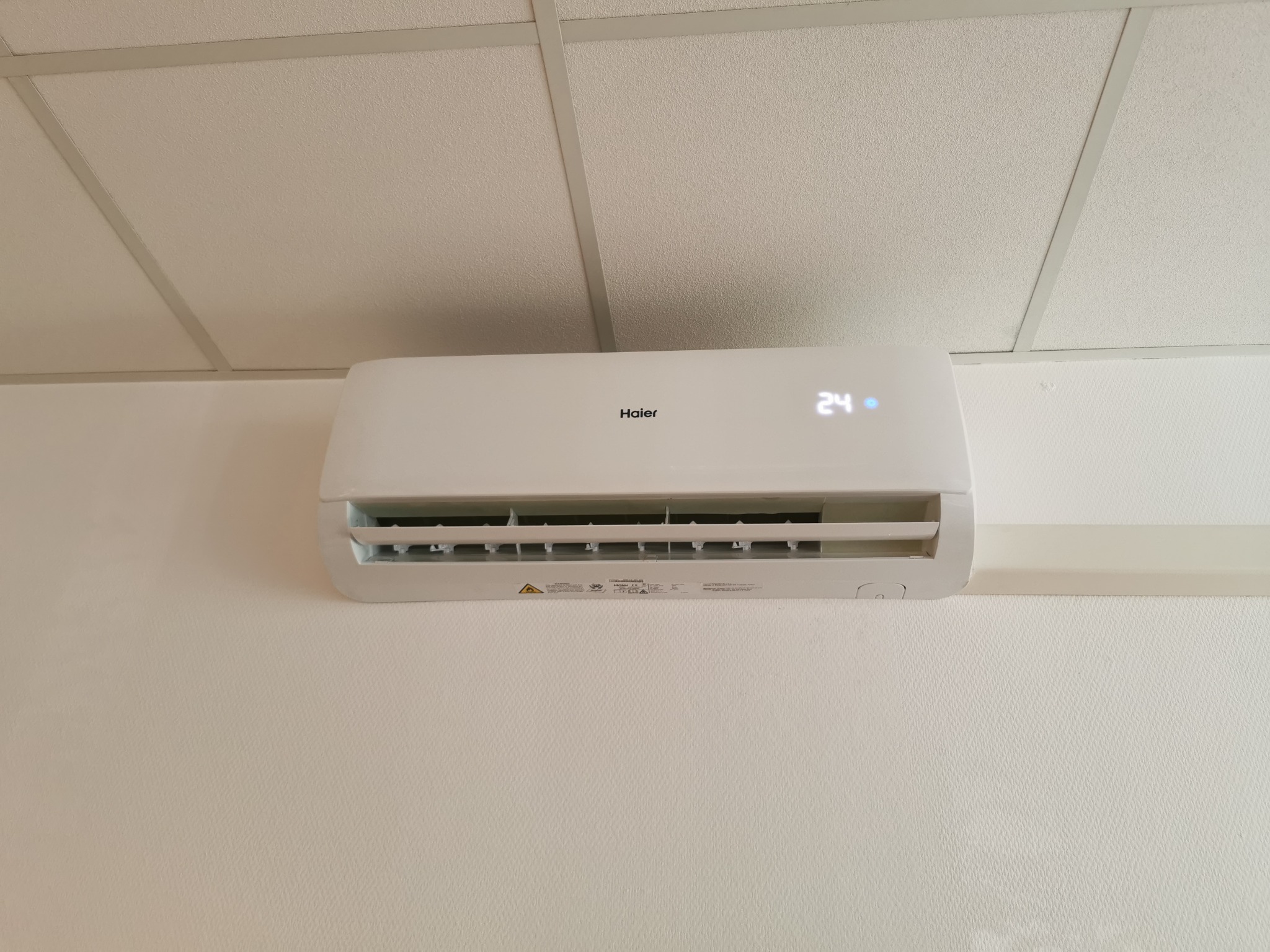 Biały klimatyzator Haier zamontowany na ścianie w pomieszczeniu biurowym, widoczny wyświetlacz z temperaturą 24 stopnie Celsjusza, sufit kasetonowy w tle.