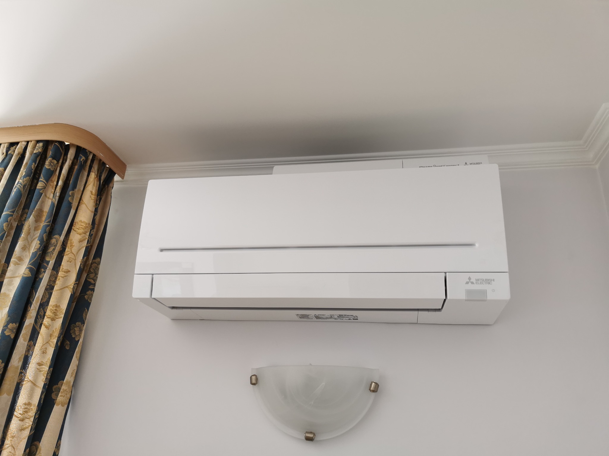 Biały klimatyzator Mitsubishi Electric zamontowany na ścianie w pomieszczeniu z ozdobnymi zasłonami i kinkietem.