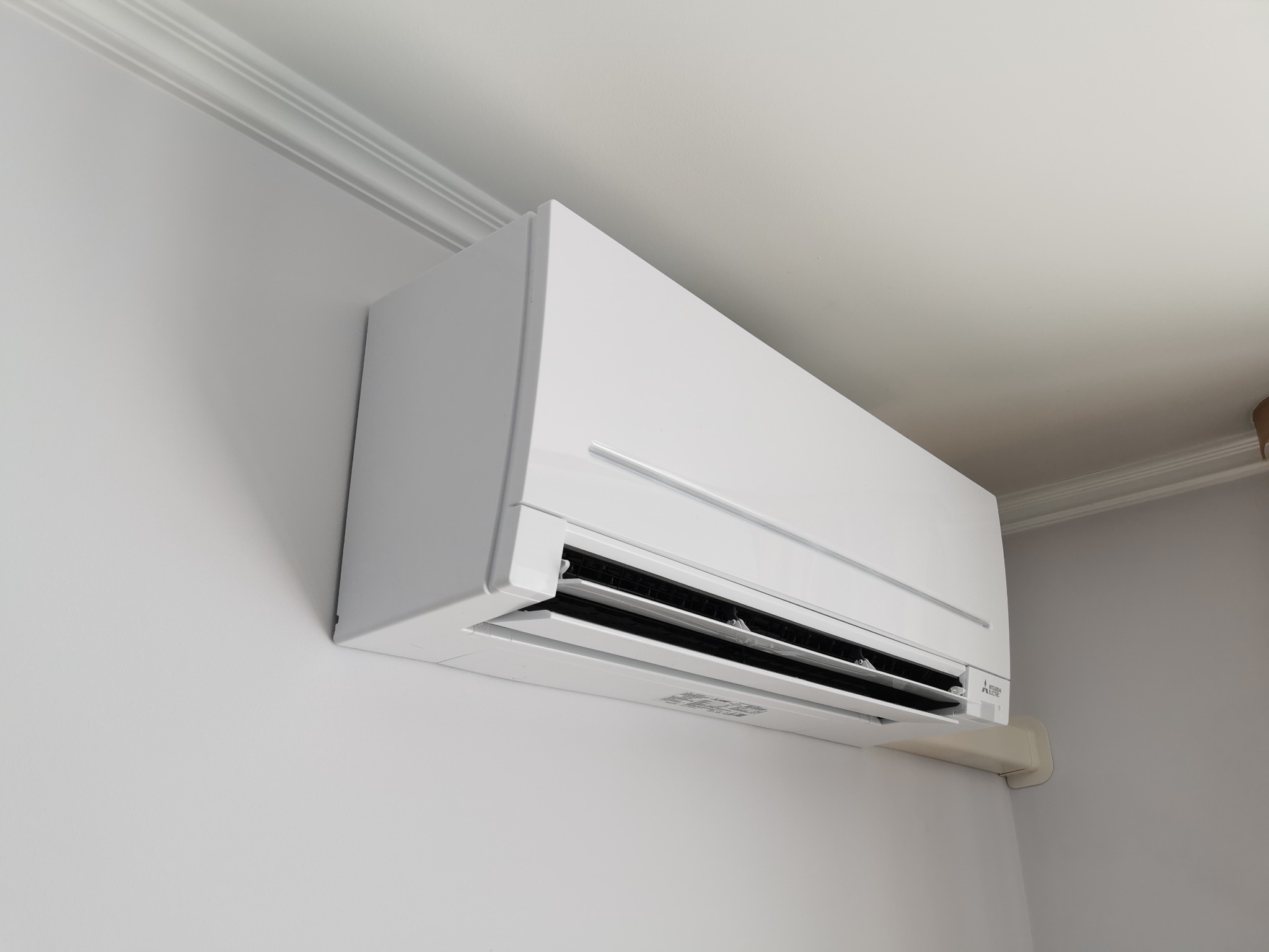 Biała jednostka wewnętrzna klimatyzatora Mitsubishi Electric zamontowana na ścianie w pomieszczeniu z białym sufitem i listwami dekoracyjnymi.