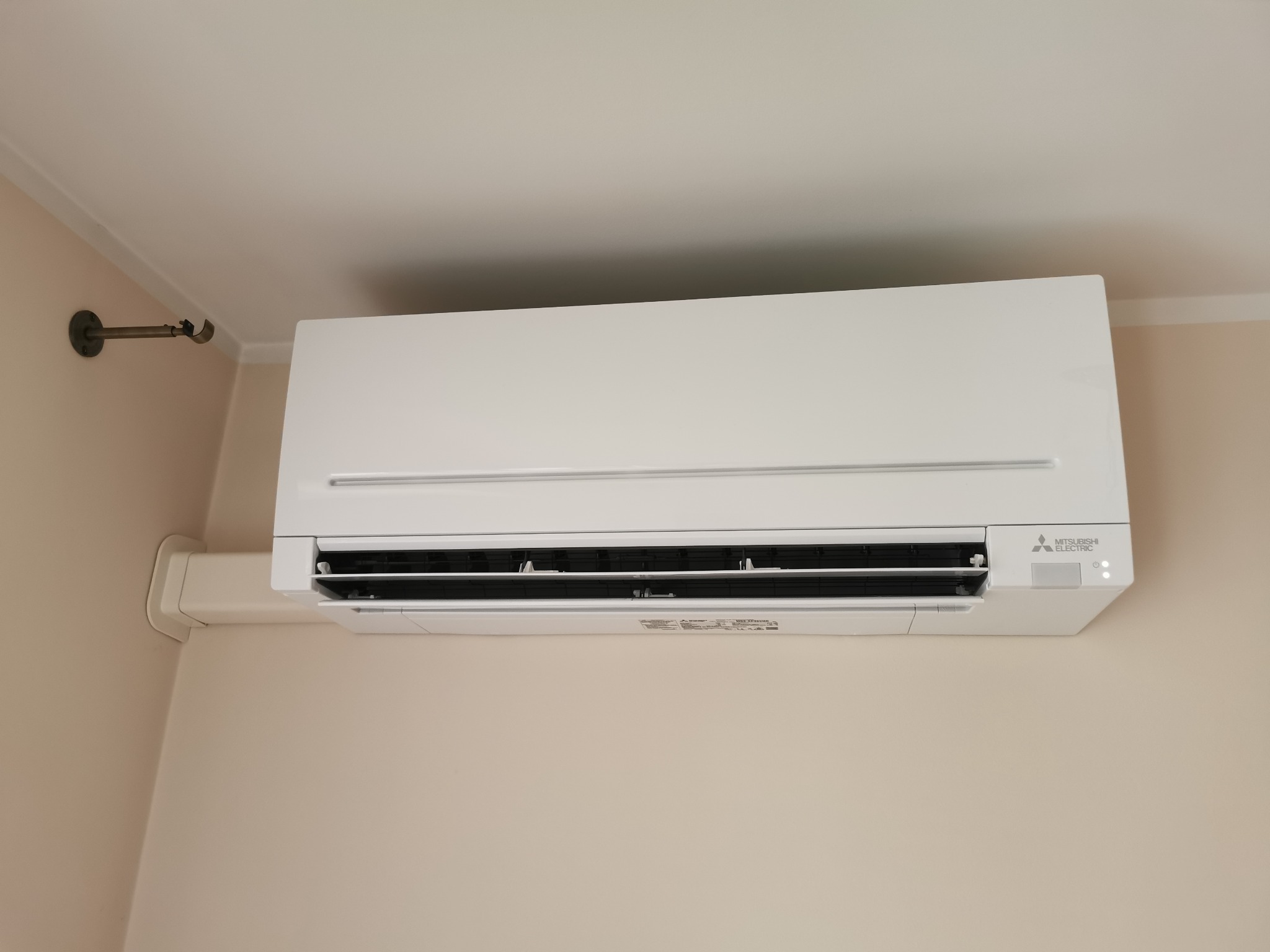 Biała jednostka wewnętrzna klimatyzatora Mitsubishi Electric zamontowana na beżowej ścianie w pomieszczeniu.