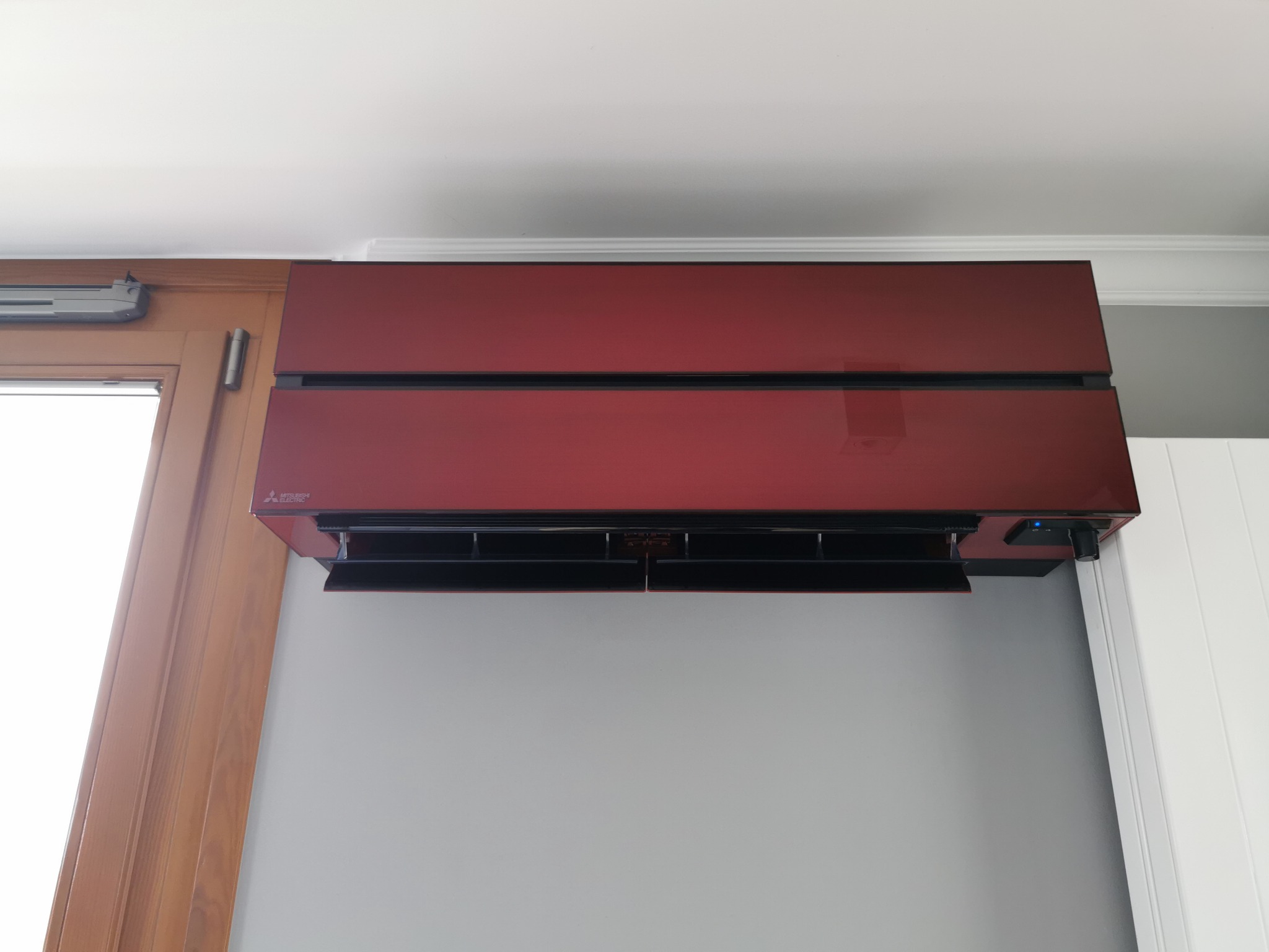 Czerwony klimatyzator Mitsubishi Electric zamontowany na szarej ścianie w pomieszczeniu z drewnianą ramą okienną i białymi panelami ściennymi.