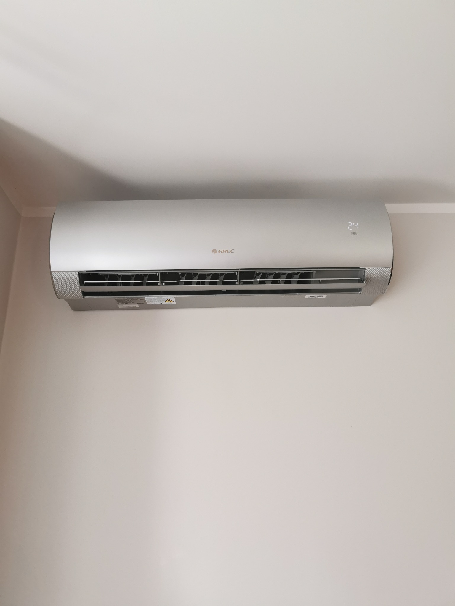 Szary klimatyzator marki Gree zamontowany na jasnej ścianie w pomieszczeniu. Na wyświetlaczu widoczna temperatura 24 stopnie Celsjusza.