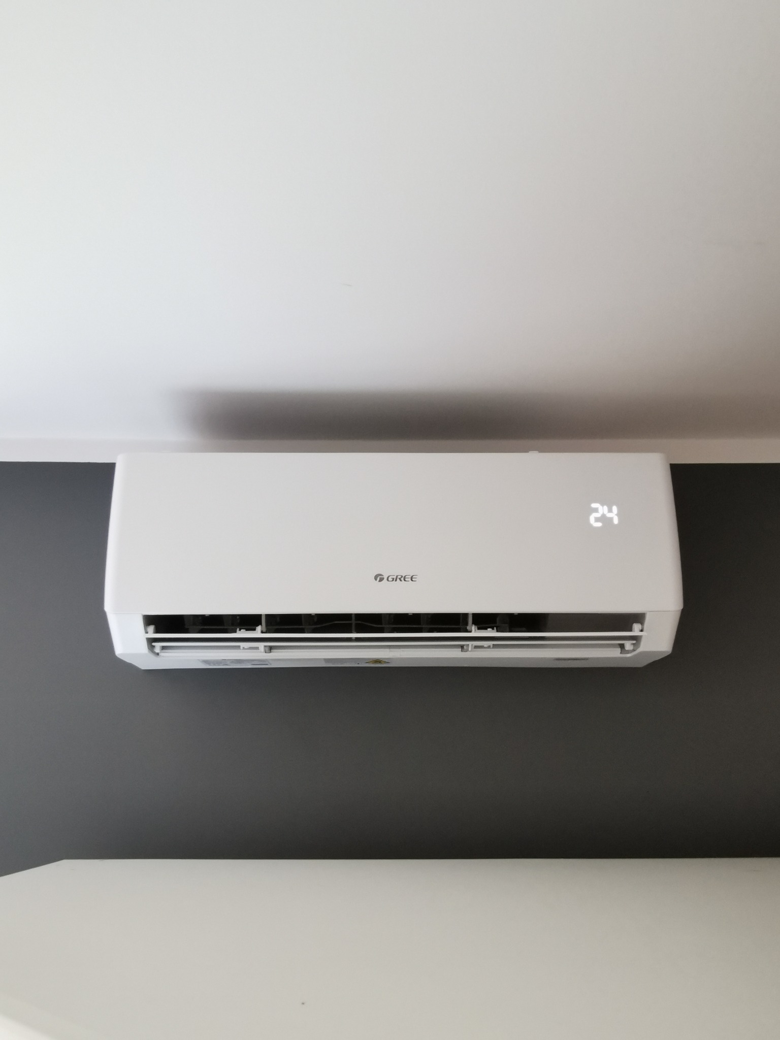 Biały klimatyzator marki Gree zamontowany na szarej ścianie, wyświetlający temperaturę 24 stopnie Celsjusza, widok z dołu.