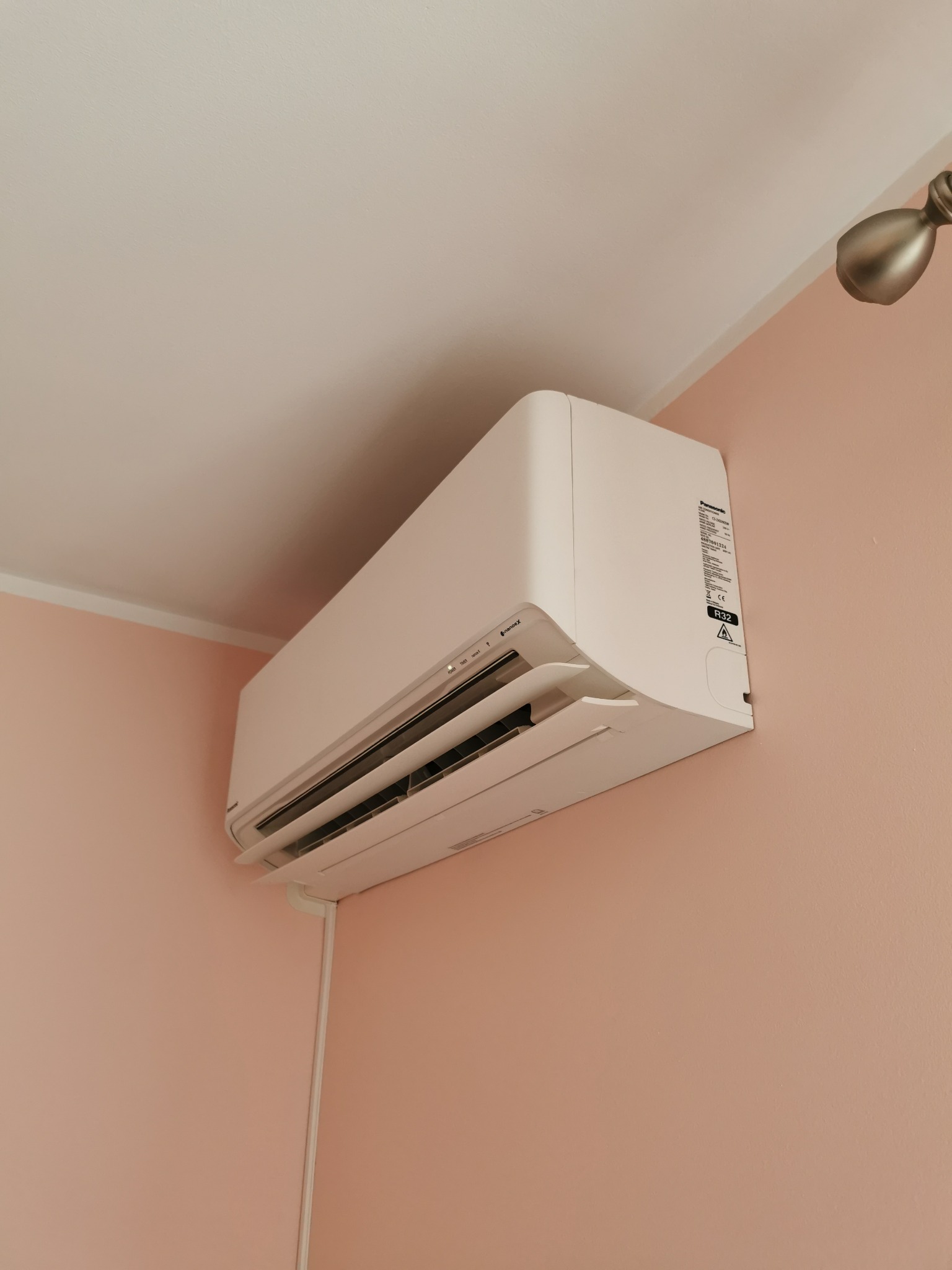 Biały klimatyzator ścienny zamontowany w rogu pokoju z różowymi ścianami, widoczne przyciski sterowania i etykieta informacyjna na boku urządzenia.