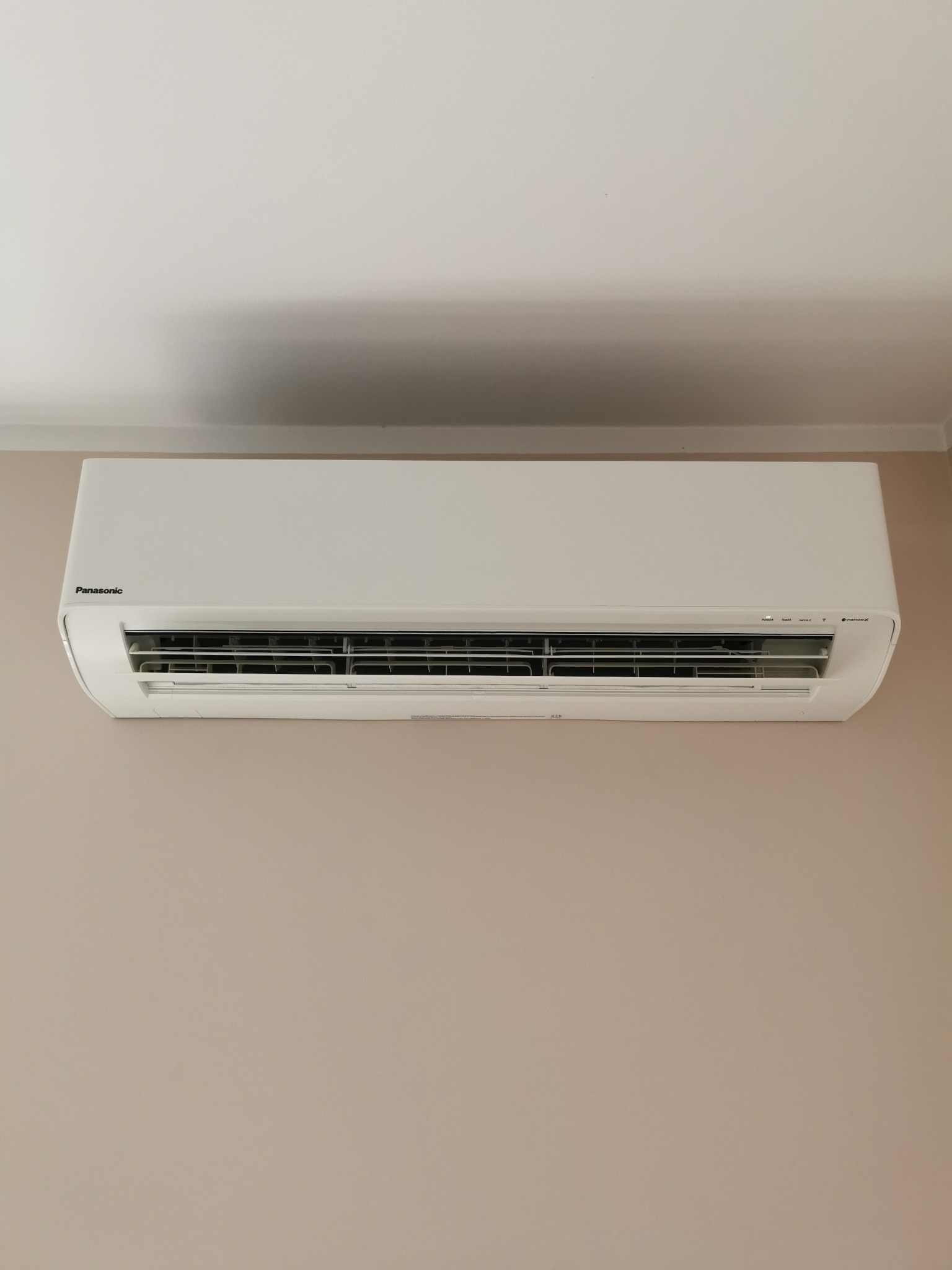 Biały klimatyzator Panasonic zamontowany na beżowej ścianie w pomieszczeniu.