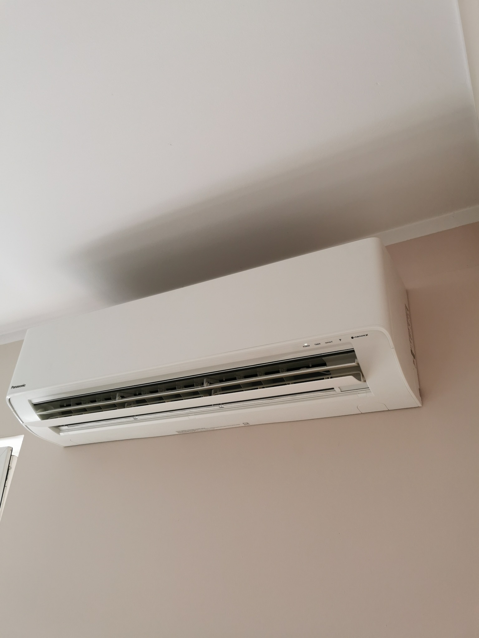 Biała jednostka wewnętrzna klimatyzacji Panasonic zamontowana na beżowej ścianie w pomieszczeniu z białym sufitem.