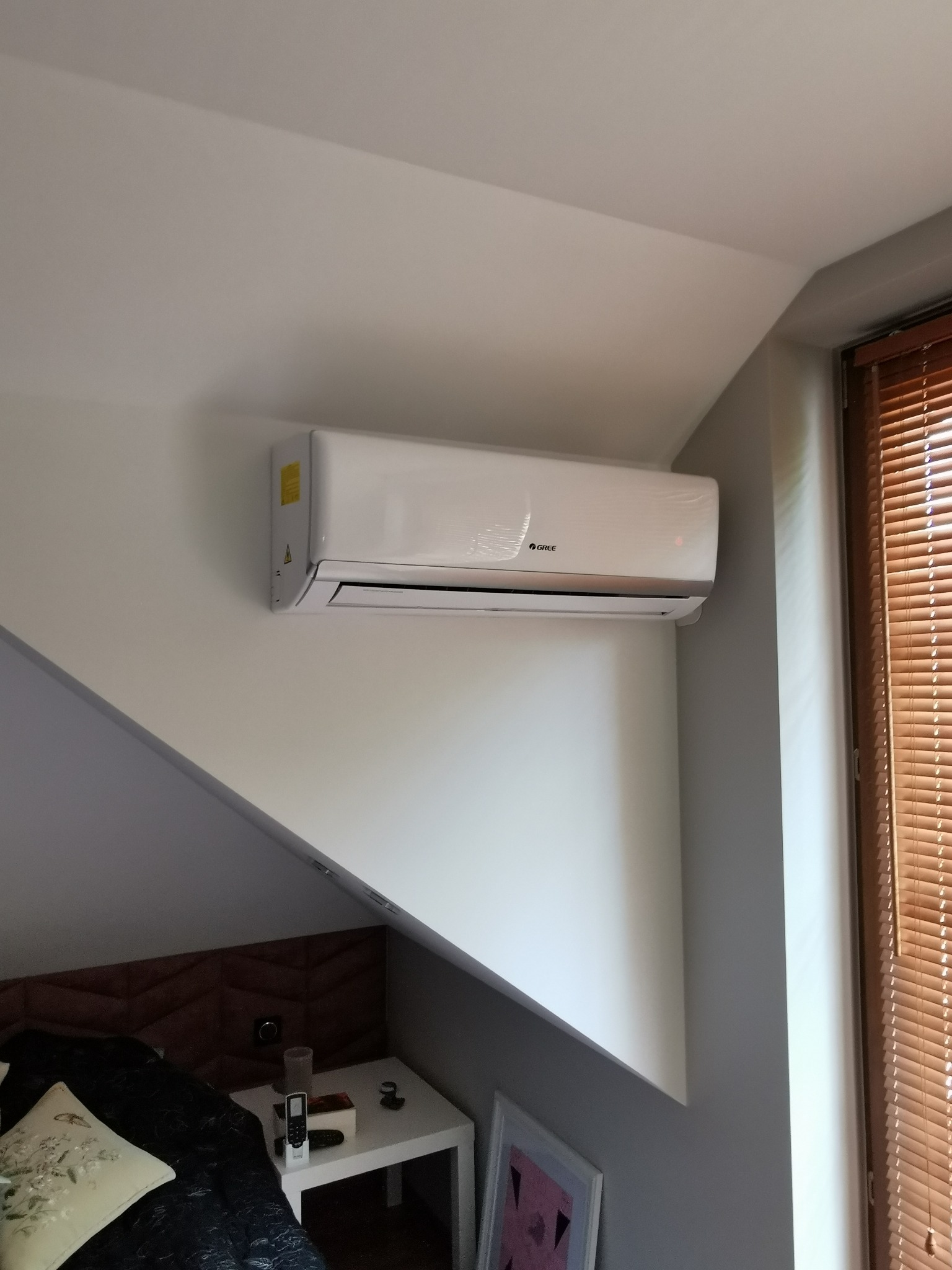 Biały klimatyzator marki Gree zamontowany na skośnej ścianie pokoju, widoczny fragment sypialni z drewnianym zagłówkiem i stolikiem nocnym, obok okno z drewnianymi żaluzjami.