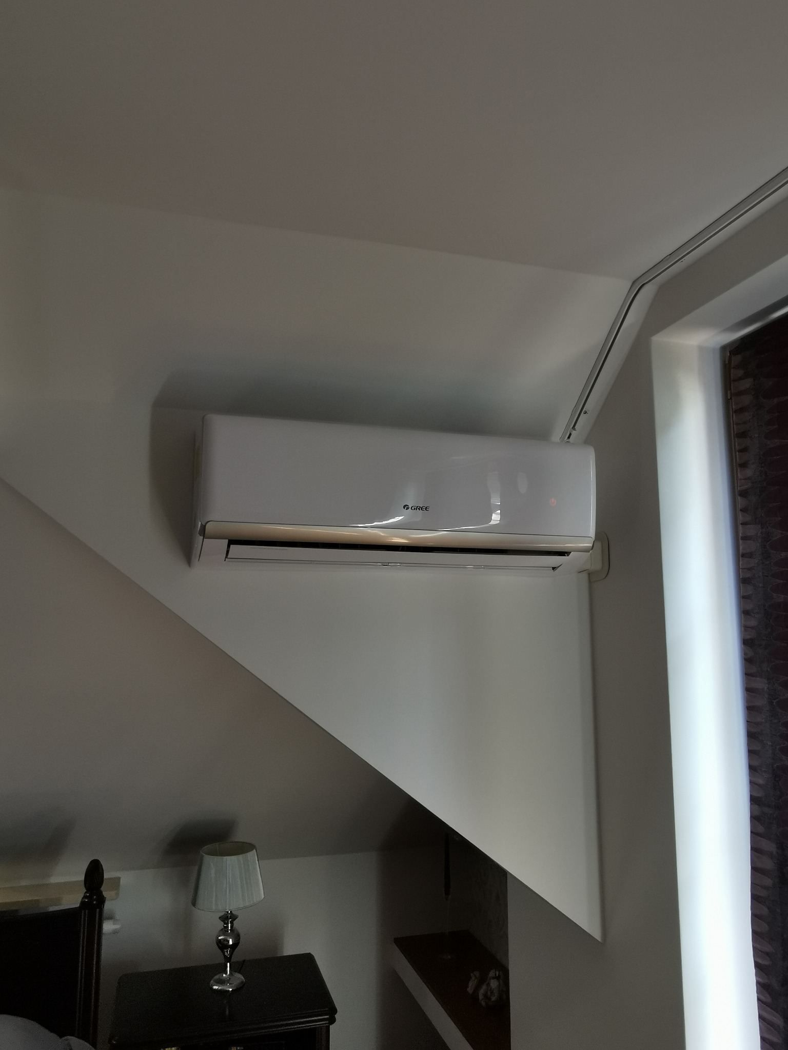 Biały klimatyzator marki Gree zamontowany na skośnej ścianie poddasza w pokoju z lampką nocną.
