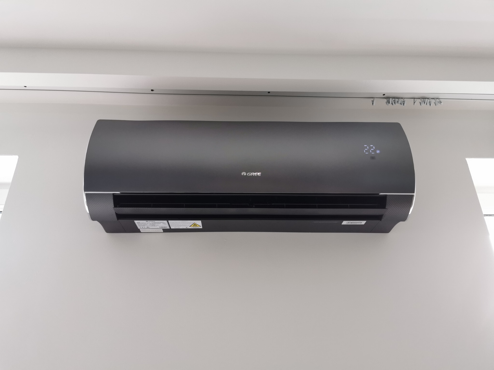 Czarny klimatyzator marki Gree zamontowany na białej ścianie, widoczne cyfry 22 na wyświetlaczu, nad urządzeniem szyna karnisza z żabkami.