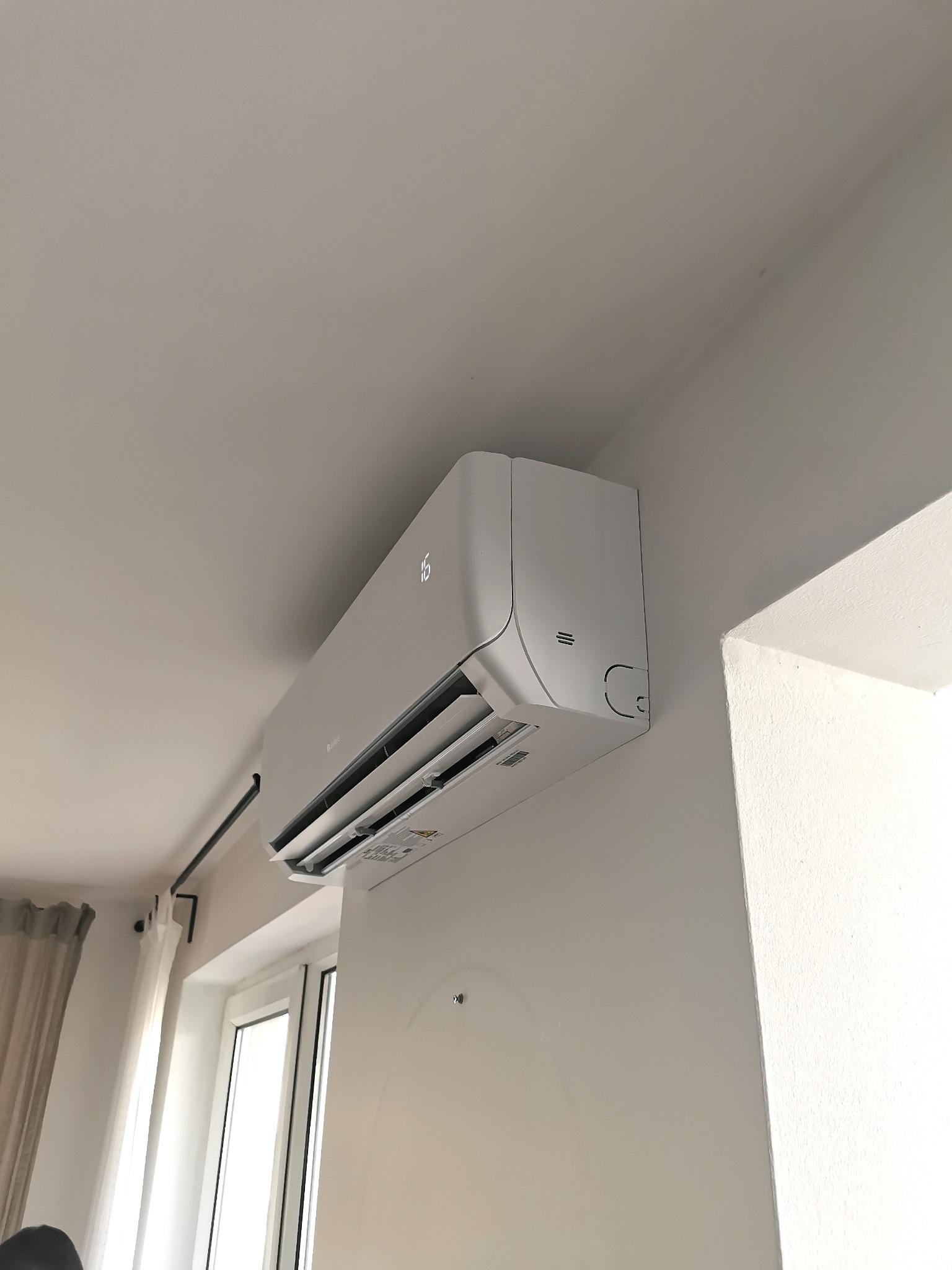 Biała klimatyzacja ścienna z wyświetlaczem temperatury '16' zamontowana w jasnym pomieszczeniu z oknem i zasłonami.