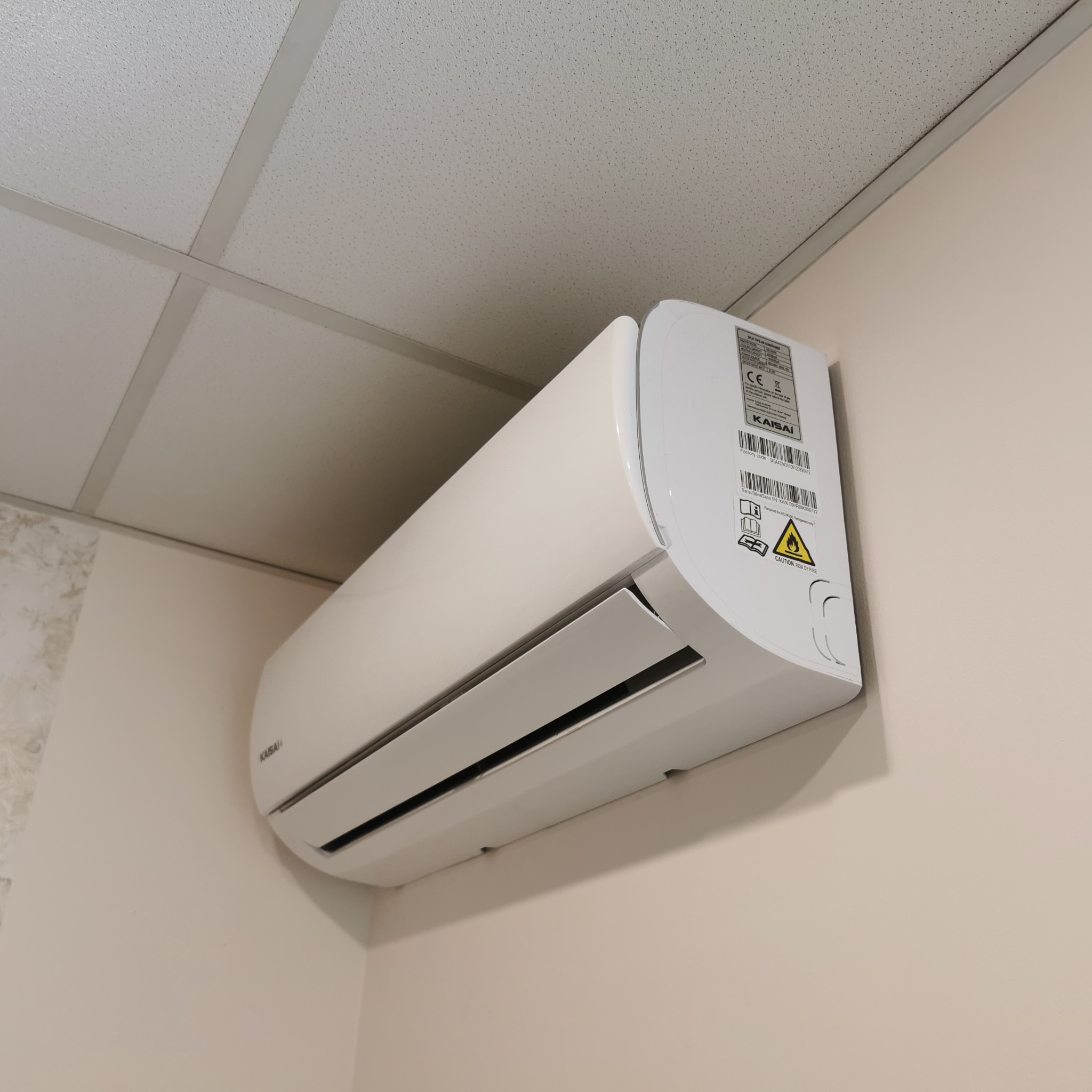 Biały klimatyzator marki Kaisai zamontowany na jasnej ścianie w pomieszczeniu z podwieszanym sufitem kasetonowym.