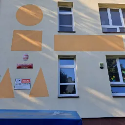 Fragment elewacji budynku przedszkola z oknami, pomalowanymi geometrycznymi kształtami i tabliczką informacyjną o monitoringu.