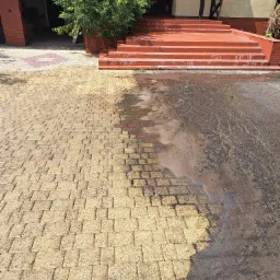 Częściowo umyta kostka brukowa przed domem z czerwonymi schodami, widoczna różnica w czystości między umytą a brudną powierzchnią.