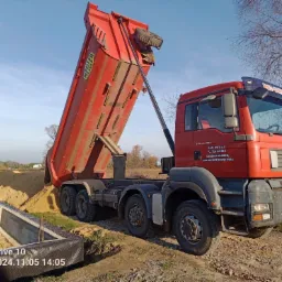 Czerwona wywrotka MAN z podniesioną skrzynią wysypuje piasek obok fundamentów domu z bloczków betonowych, zabezpieczonych folią kubełkową.