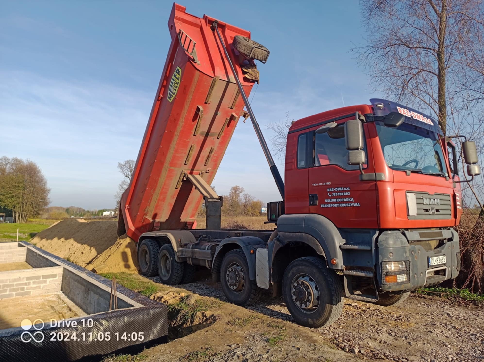 Czerwona wywrotka MAN z podniesioną skrzynią wysypuje piasek obok fundamentów domu z bloczków betonowych, zabezpieczonych folią kubełkową.