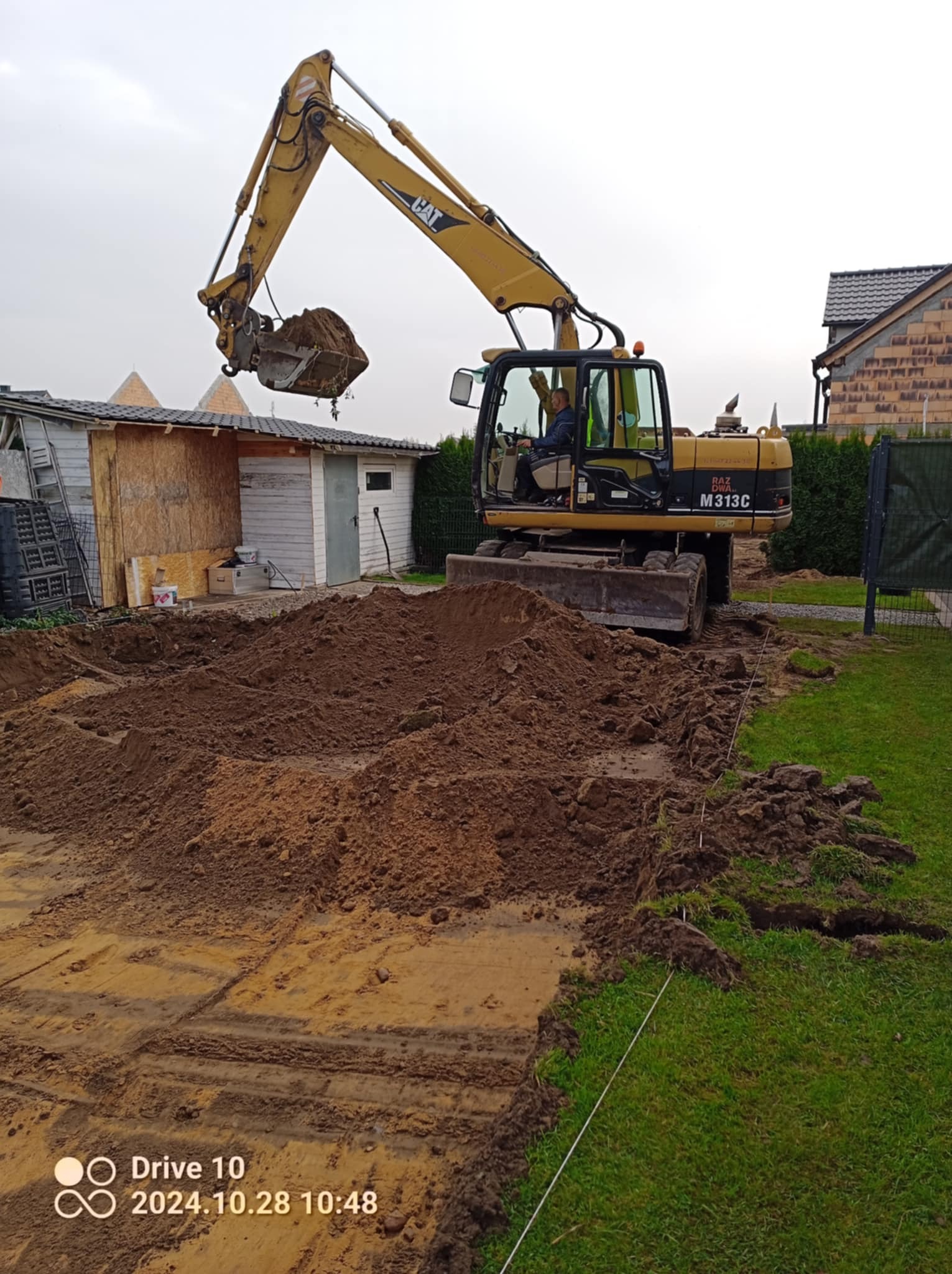 Żółta koparka CAT M313C podczas prac ziemnych na działce, widoczne wyznaczone linie pod fundamenty, operator w kabinie, w tle budynek gospodarczy i dom.