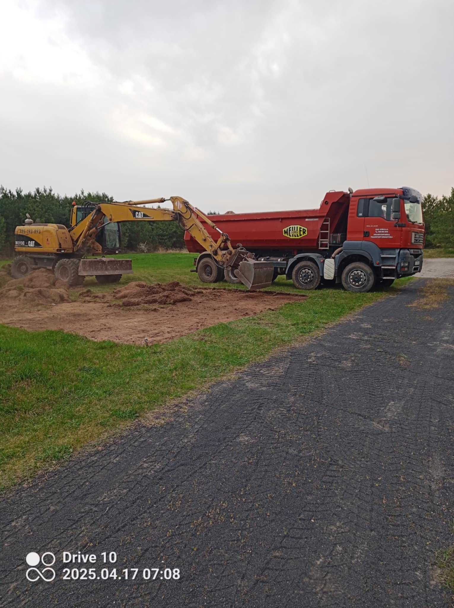 Żółta koparka CAT ładuje ziemię do czerwonej ciężarówki Meiller Kipper na tle zieleni i pochmurnego nieba. Prace ziemne w toku.
