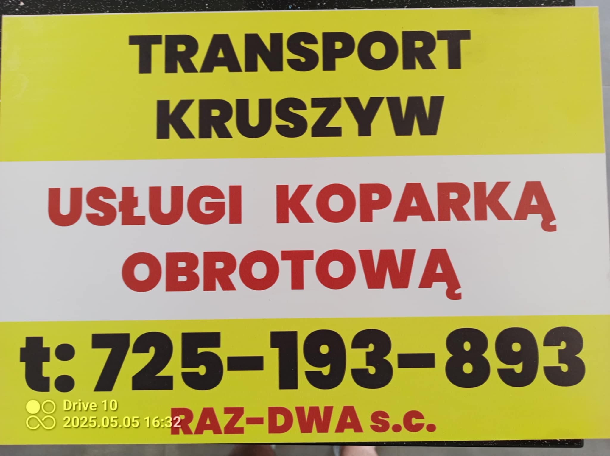Żółto-biała tablica informacyjna z czarnym napisem 'TRANSPORT KRUSZYW' i czerwonym 'USŁUGI KOPARKĄ OBROTOWĄ', poniżej numer telefonu.