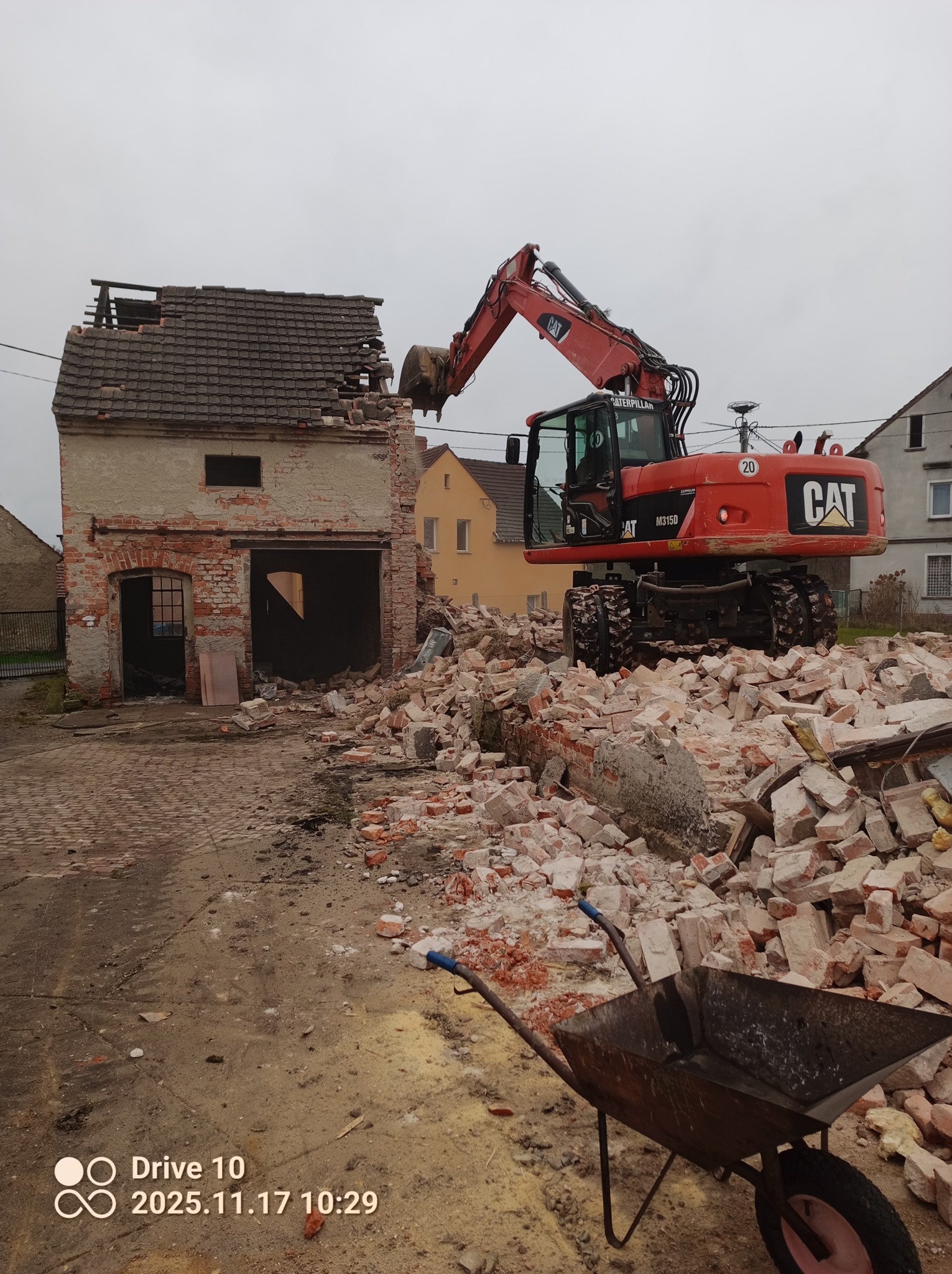 Czerwona koparka CAT M315D burzy ceglany budynek. Gruz i taczka na pierwszym planie. Widoczne fragmenty dachu i ścian. Pochmurne niebo w tle.