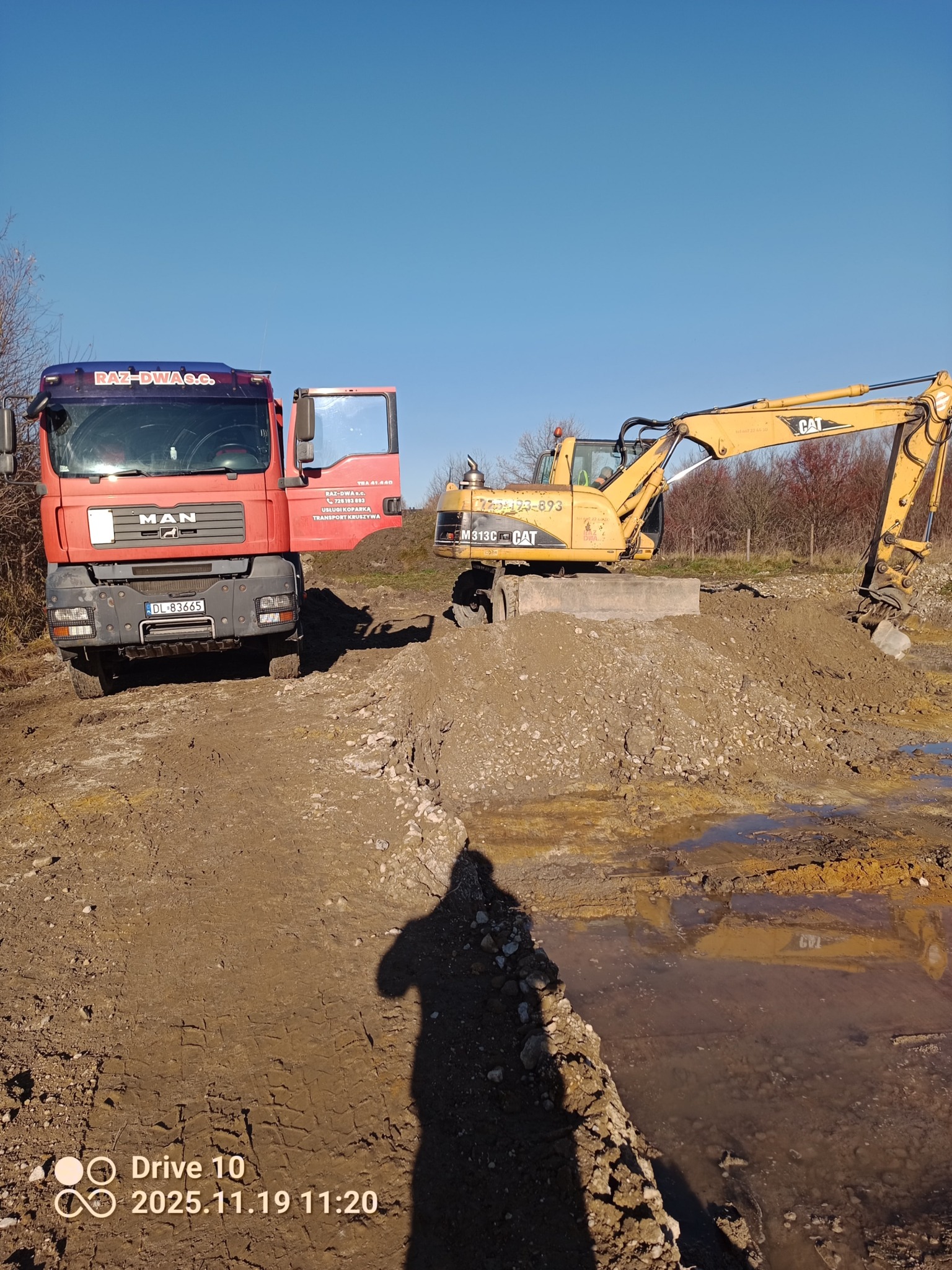 Czerwona ciężarówka MAN z otwartymi drzwiami obok żółtej koparki CAT na placu budowy z ziemią i kałużami. Widoczny cień osoby. Błękitne niebo w tle.