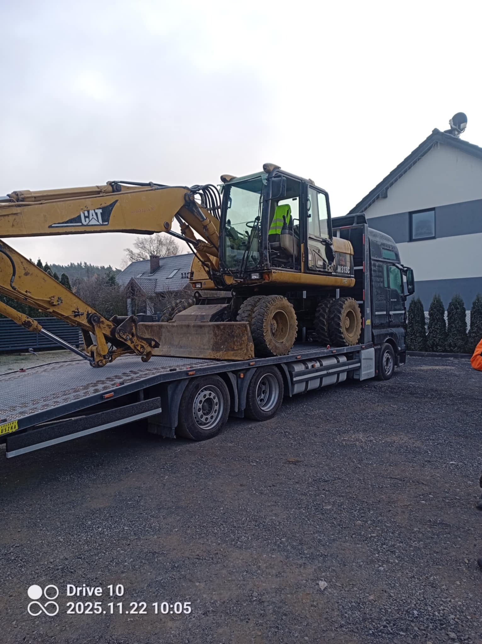 Żółta koparka CAT M313C na lawecie ciężarówki na żwirowym podłożu, z budynkiem w tle. Transport maszyny budowlanej.