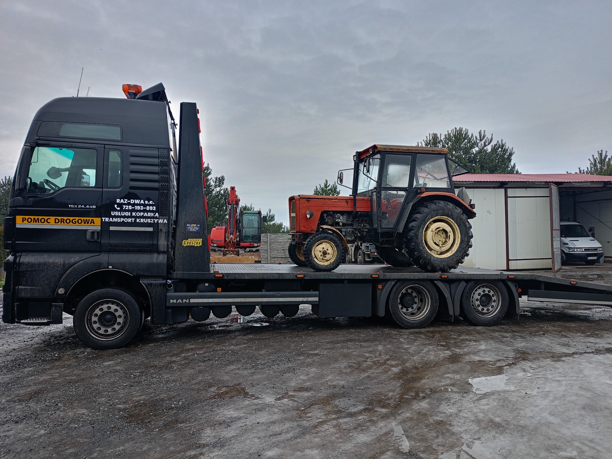 Transport niskopodwoziowy w Legnicy: Czarna laweta MAN z czerwonym traktorem na platformie. W tle widoczny sprzęt budowlany i budynek gospodarczy.