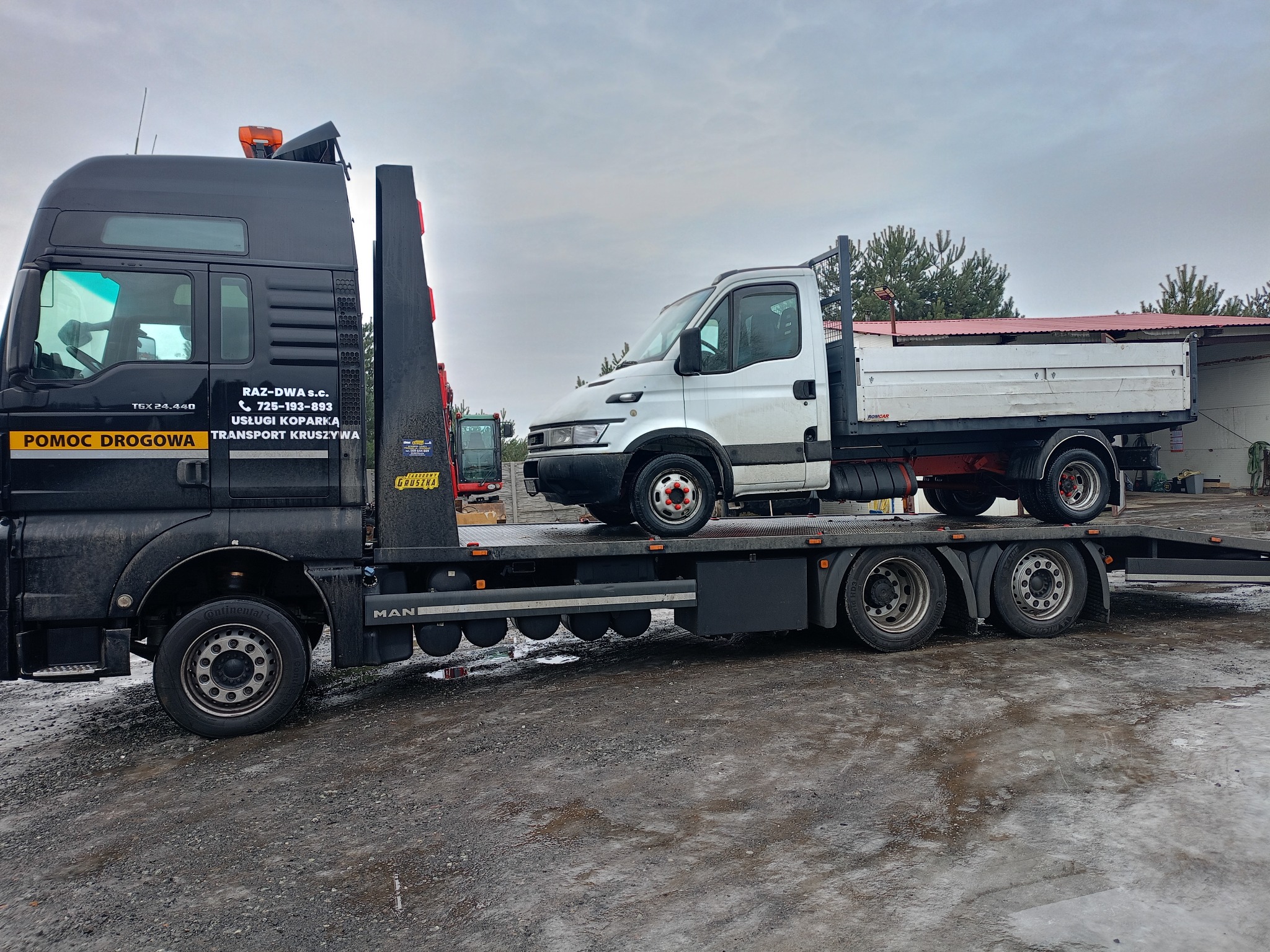 Czarna laweta MAN z białym samochodem dostawczym na platformie. Widoczne logo firmy 'POMOC DROGOWA' i 'TRANSPORT KRUSZYWA'.