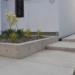 Betonowa ścieżka i schody prowadzące wzdłuż białego budynku w Dębnie, z rabatą kwiatową w betonowej obudowie po lewej stronie.