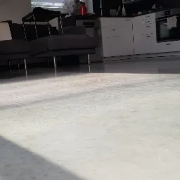 Jasna, gładka posadzka w salonie z szarym narożnikiem na metalowych nogach i białą zabudową kuchenną w tle, oświetlona naturalnym światłem