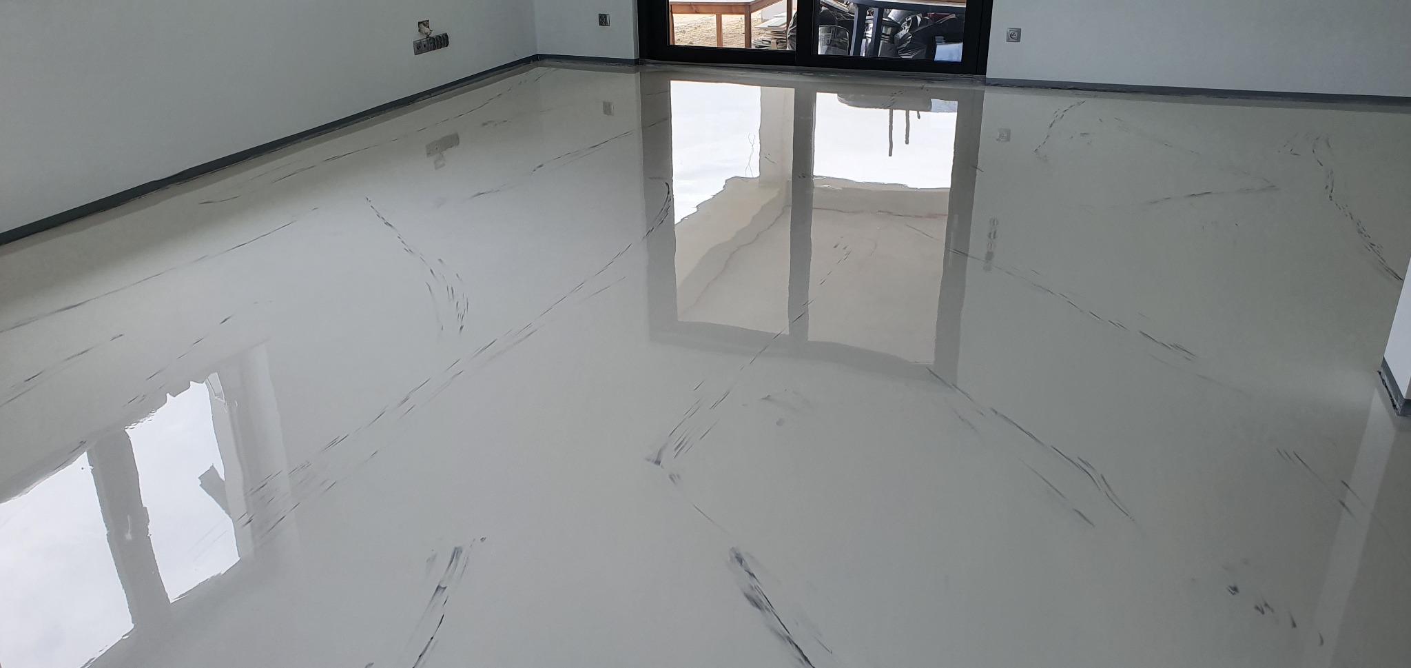 Błyszcząca, jasnoszara posadzka żywiczna z ciemnymi żyłkami imitującymi marmur, odbijająca światło z okna balkonowego.