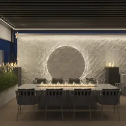 Zadaszony taras z długim stołem i paleniskiem, krzesła z plecionki, ściana z kamienia z okrągłym podświetleniem, dekoracyjne trawy i płomienie w szklanych obudowach.