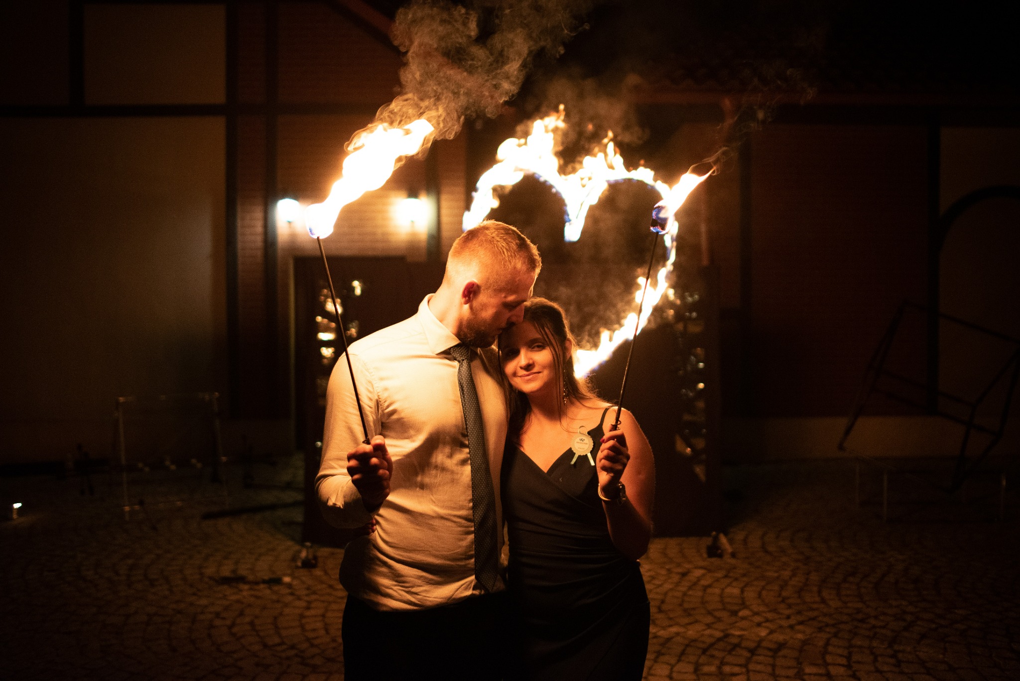 Para trzymająca płonące pochodnie w kształcie serca podczas pokazu pirotechnicznego na tle budynku z cegły.