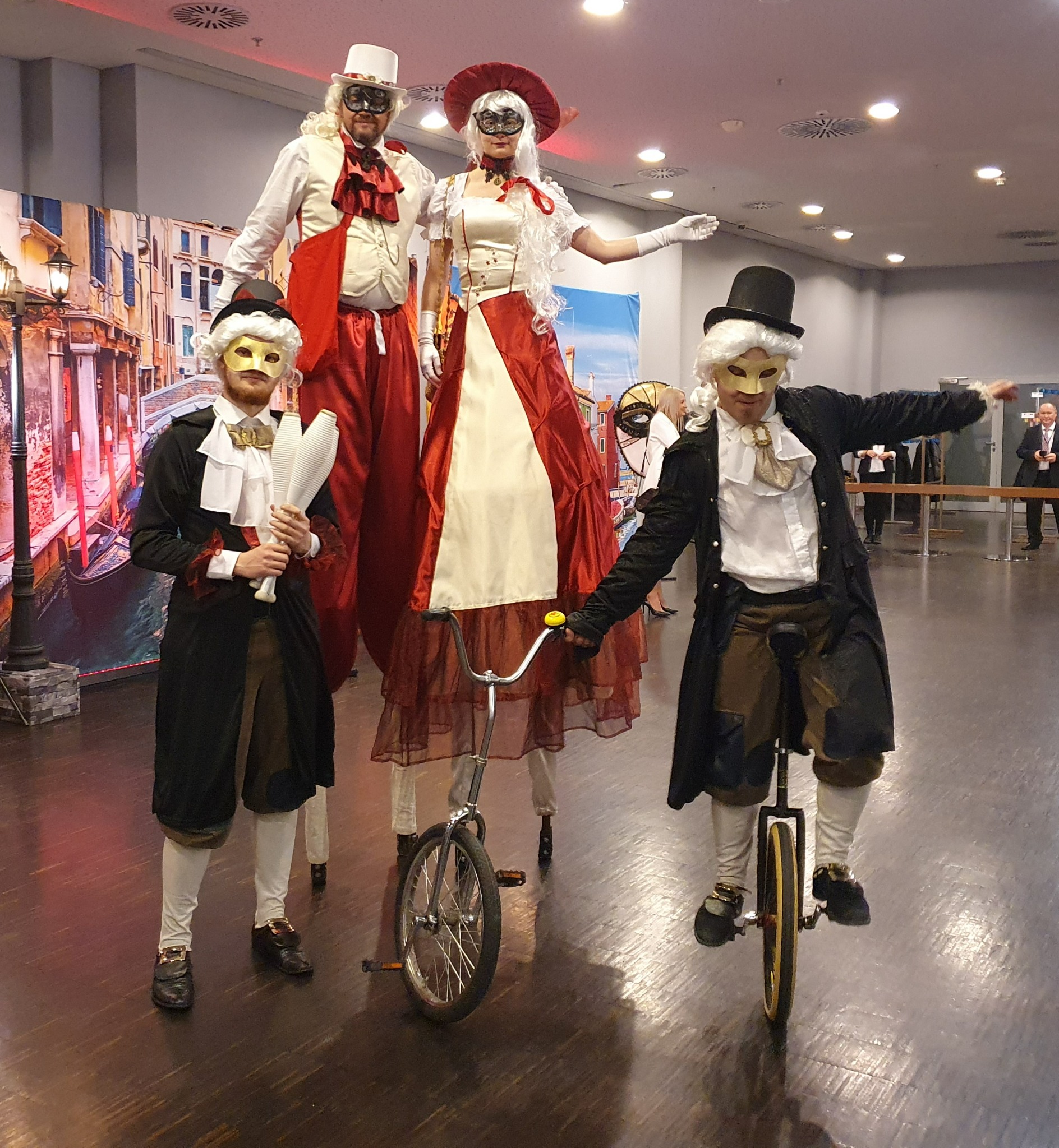Grupa czterech osób w kostiumach weneckich, w tym dwie na szczudłach i jedna na monocyklu, pozują na tle dekoracji z motywem weneckim.