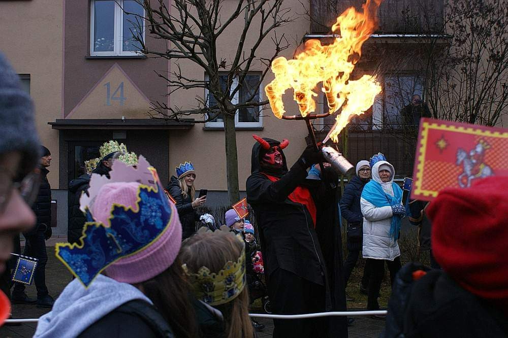Uliczna parada z osobą przebraną za diabła, trzymającą płonący rekwizyt, otoczoną przez ludzi w koronach i zimowych ubraniach, na tle budynku.
