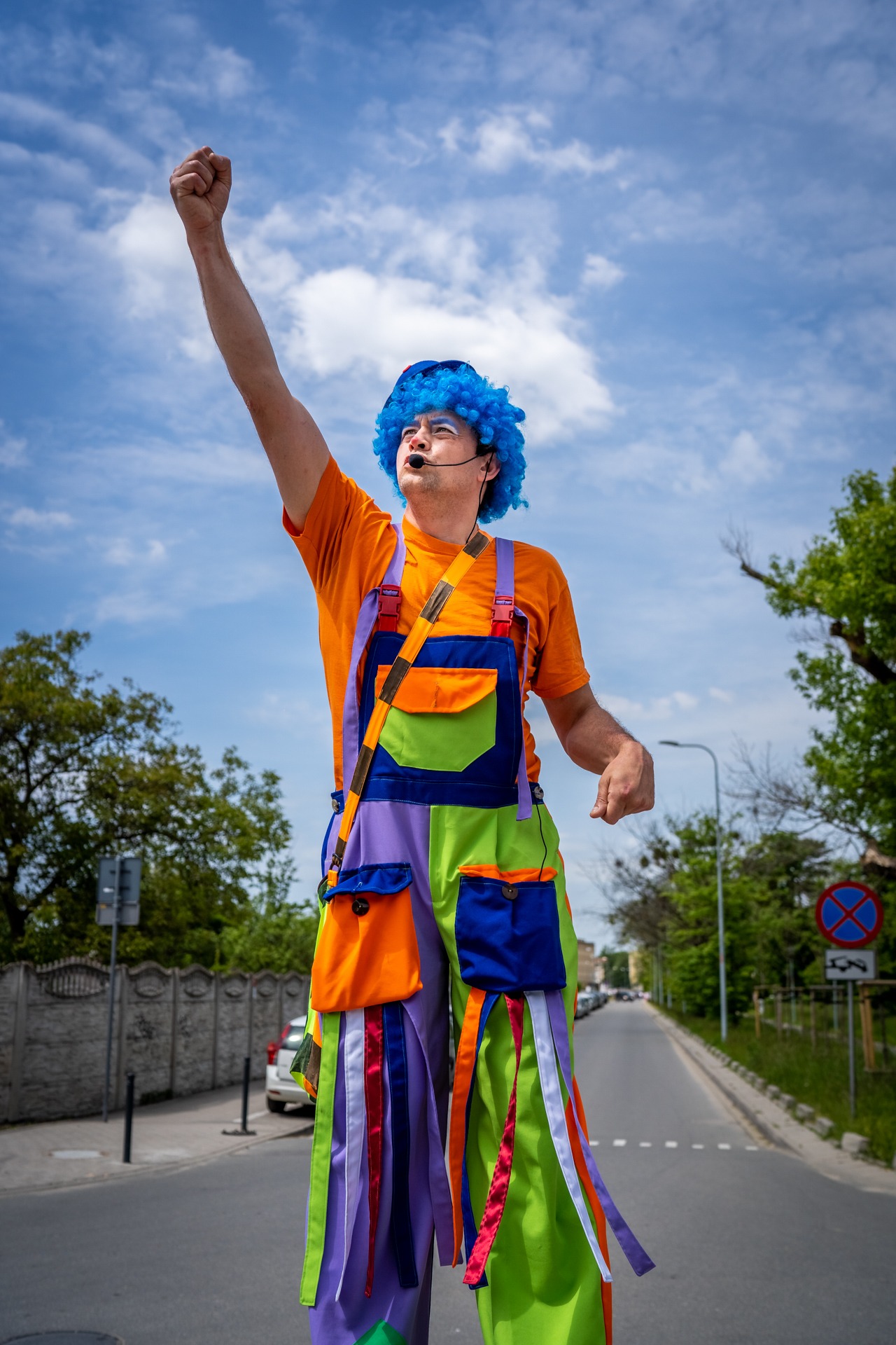 Uśmiechnięty mężczyzna w kostiumie klauna, z niebieską peruką i mikrofonem nagłownym, stojący na ulicy z uniesioną pięścią w geście triumfu.