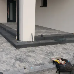 Ułożona kostka brukowa w odcieniach szarości prowadząca do schodów wejściowych. Na pierwszym planie widoczny plecak z rękawicami roboczymi, a w tle narzędzia i akcesoria brukarskie.