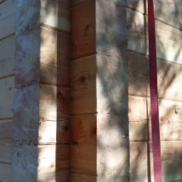 Fragment ściany domu z bali o grubości 24cm z modrzewia polskiego, widoczne łączenia bali i czerwony pas transportowy.