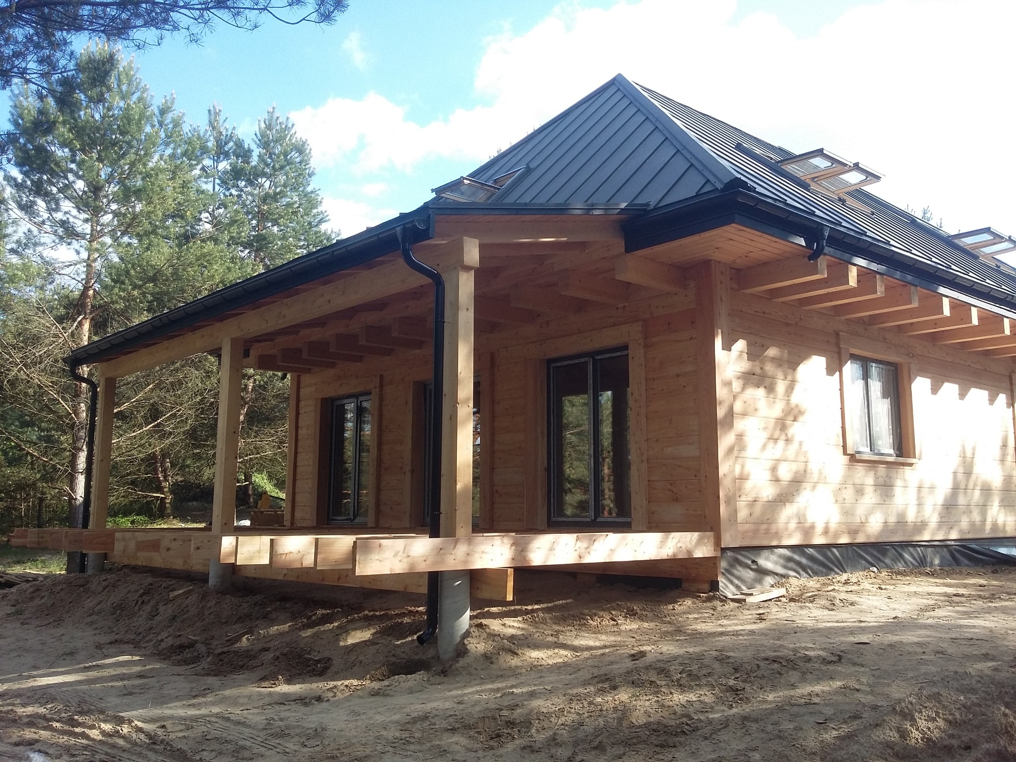 Surowy, drewniany dom szkieletowy z modrzewiową elewacją w trakcie budowy, widoczne czarne okna, metalowy dach z lukarnami i częściowo wykonany taras, otoczony piaszczystym terenem i drzewami.