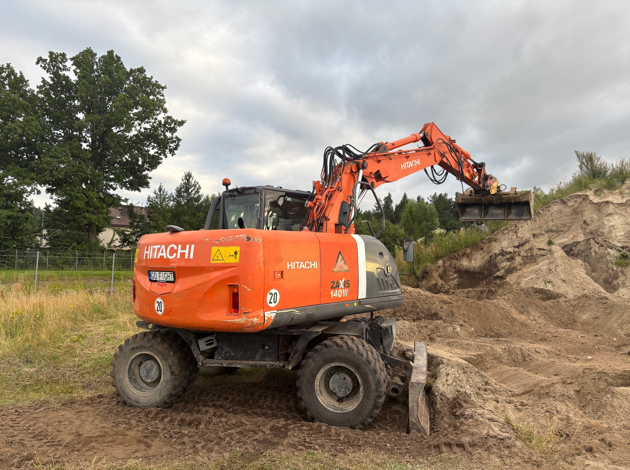 Pomarańczowa koparka kołowa Hitachi ZX140W nabiera ziemię z pryzmy na placu budowy, w tle drzewa i pochmurne niebo.