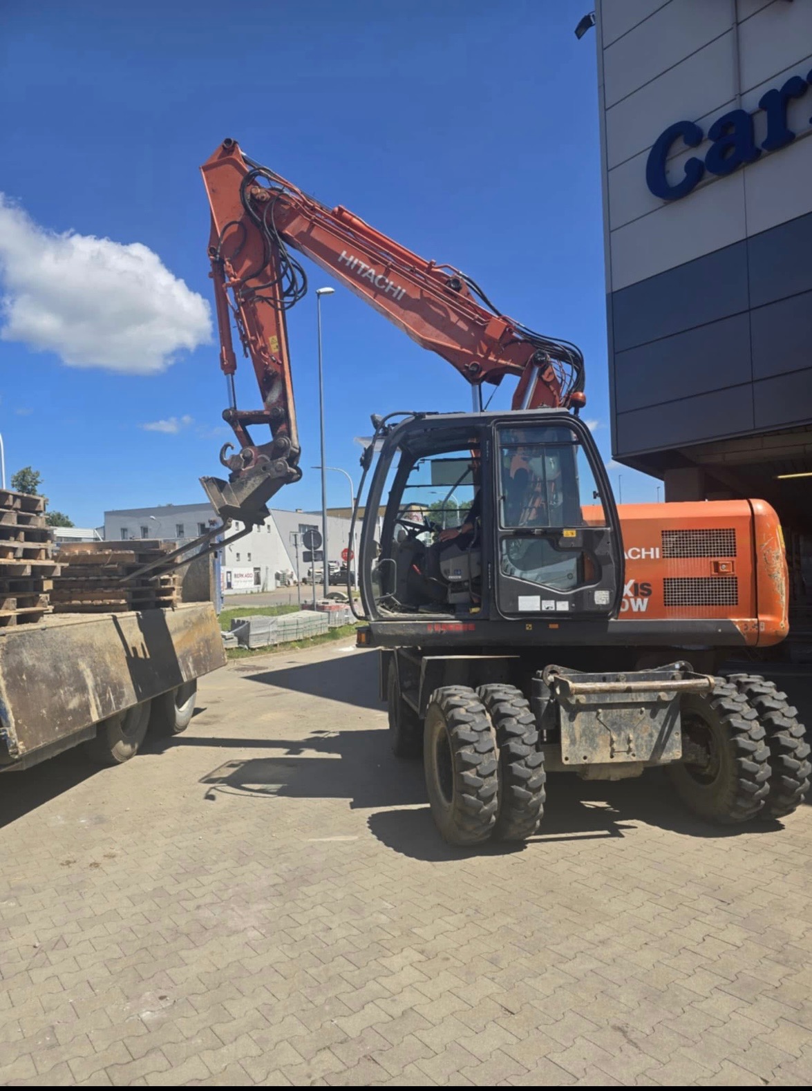 Pomarańczowa koparka Hitachi ładuje palety na przyczepę na tle błękitnego nieba z chmurami i budynkiem Carrefour. Operator w kabinie.