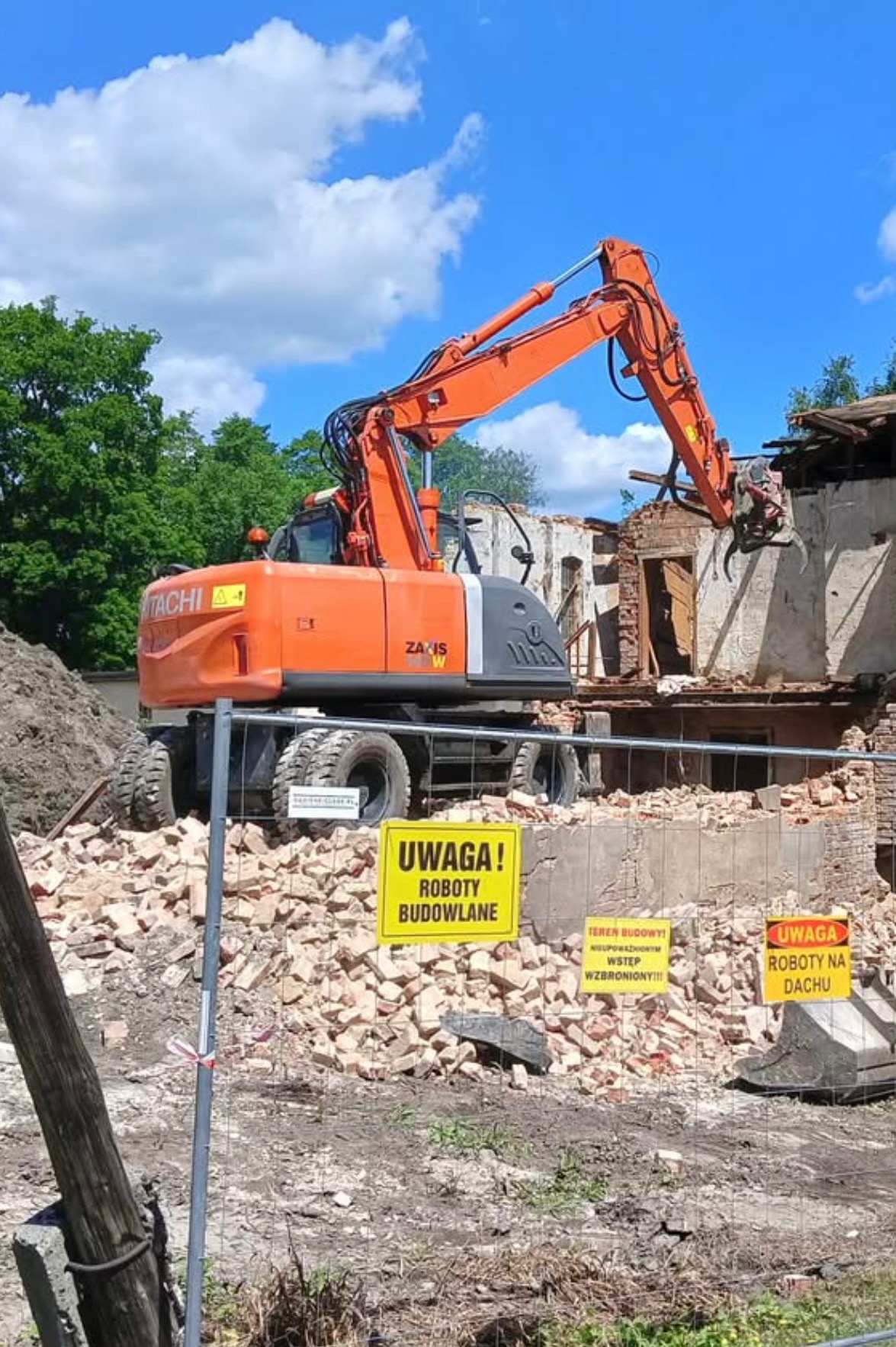 Pomarańczowa koparka Hitachi ZAXIS 145W na tle gruzów i fragmentów budynku w trakcie rozbiórki. Żółte tablice ostrzegawcze informują o robotach budowlanych.