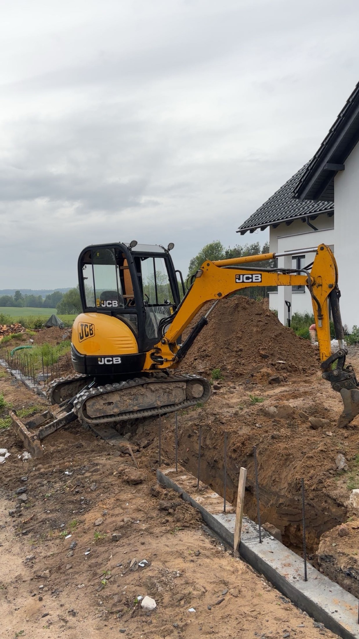 Żółta koparka JCB przy wykopie fundamentów z częściowo zalanym betonem i wystającymi prętami zbrojeniowymi na tle domu i zieleni.