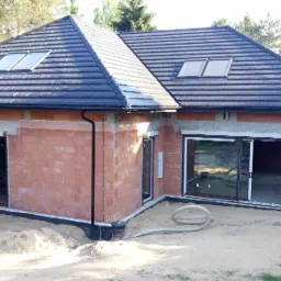 Budowa domu jednorodzinnego w stanie surowym zamkniętym, widoczne okna, drzwi przesuwne, dachówka ceramiczna i częściowo ocieplone ściany z czerwonej cegły, na zewnątrz piasek i rury wentylacyjne.