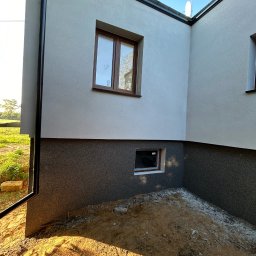 Nidecki Elewacje - Fragment domu z ocieploną elewacją, widoczna dwukolorowa struktura tynku, okna z brązowymi ramami i rynna spustowa przy narożniku budynku, teren wokół budynku częściowo przygotowany...