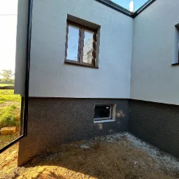 Fragment domu z ocieploną elewacją, widoczna dwukolorowa struktura tynku, okna z brązowymi ramami i rynna spustowa przy narożniku budynku, teren wokół budynku częściowo przygotowany...