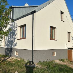 Nidecki Elewacje - Dwukondygnacyjny dom jednorodzinny z jasną elewacją i szarą podmurówką, brązowe ramy okienne, metalowy dach, widoczny fragment ogrodu i cień drzewa.