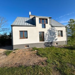 Nidecki Elewacje - Nowoczesny dom z białą elewacją i szarym dachem blachodachówką, widoczny z poziomu gruntu, z fragmentem niezagospodarowanego terenu wokół, w słoneczny dzień.