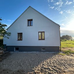 Nidecki Elewacje - Świeżo otynkowana, biała elewacja domu jednorodzinnego z brązowymi oknami, widok z poziomu gruntu na tle błękitnego nieba z chmurami.